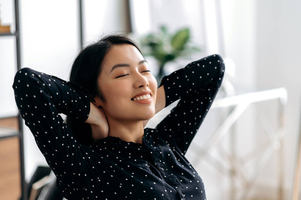 Pretty, asian girl resting during work. Positive female manager or freelancer, sits at office, takes a break from work, with eyes closed, throwing his hands behind his head, smiles, dreams of rest - Photo, Image