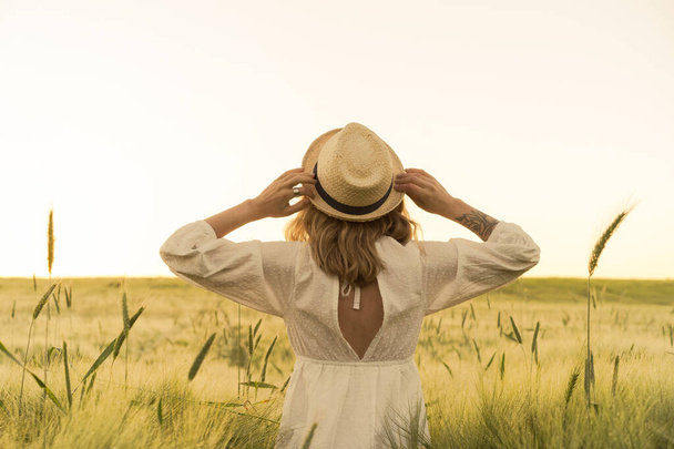 young beautiful woman with blond long hair in a white dress in a straw hat on a wheat field. Flying hair in the sun, summer. Time for dreamers, golden sunset. - Foto, afbeelding