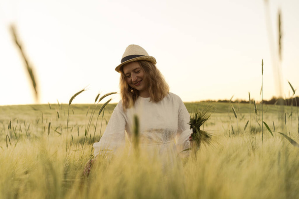 young beautiful woman with blond long hair in a white dress in a straw hat collects bouquet on a wheat field. Flying hair in the sun, summer. Time for dreamers, golden sunset. - Фото, зображення