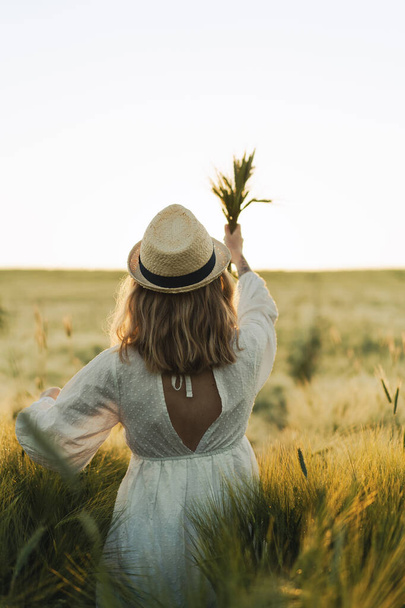 young beautiful woman with blond long hair in a white dress in a straw hat on a wheat field. Flying hair in the sun, summer. Time for dreamers, golden sunset. - Fotó, kép