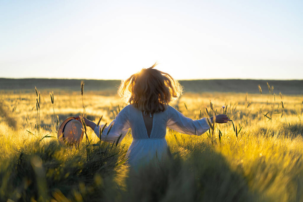 young beautiful woman with long blond hair in a white dress on a wheat field in the early morning at sunrise. Summer is the time for dreamers, flying hair, a woman running across the field in the rays of the sun. travels - Фото, изображение
