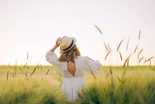 金髪の若い美女が麦畑の麦藁帽子の白いドレスを着た。太陽の下で飛行髪、夏。夢の時間黄金の夕日. - 写真・画像