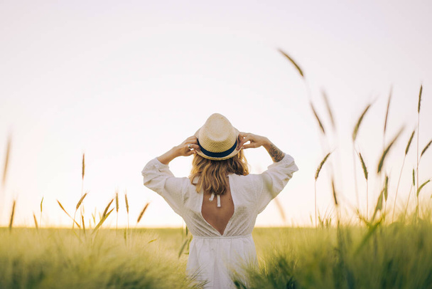 young beautiful woman with blond long hair in a white dress in a straw hat on a wheat field. Flying hair in the sun, summer. Time for dreamers, golden sunset. - 写真・画像