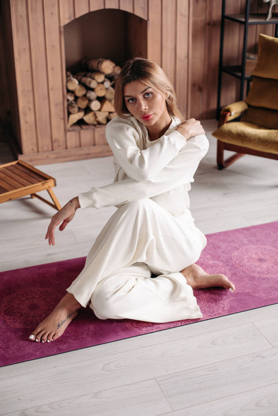 Belle jeune fille portant des vêtements de sport naturels écologiques en coton blanc, assise sur un tapis dans un intérieur confortable à la maison - Photo, image