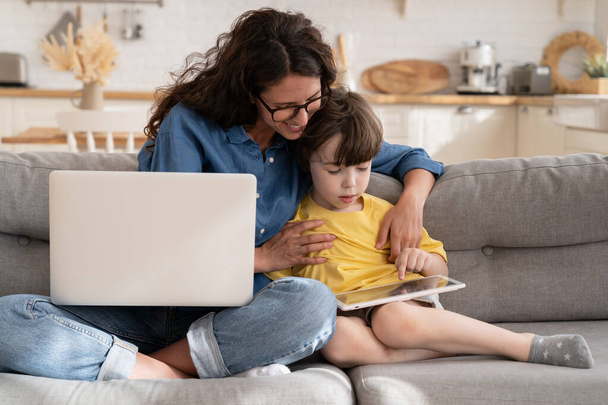 Η μαμά και το παιδί γιος χρήση tablet κάθονται στον καναπέ, χαμογελαστή μητέρα βοηθήσει το παιδί με e-learning μαθήματα ή παιχνίδια - Φωτογραφία, εικόνα