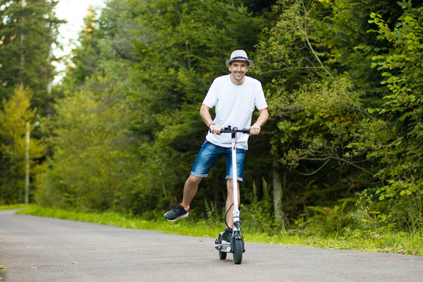 Elektrikli taşıma. Beyaz tişörtlü, güneş gözlüklü ve şapkalı genç yakışıklı adam güneşli bir yaz gününde elektrikli scooter bisikletini sürmeye hazır. Ekolojik ulaşım kavramı. - Fotoğraf, Görsel