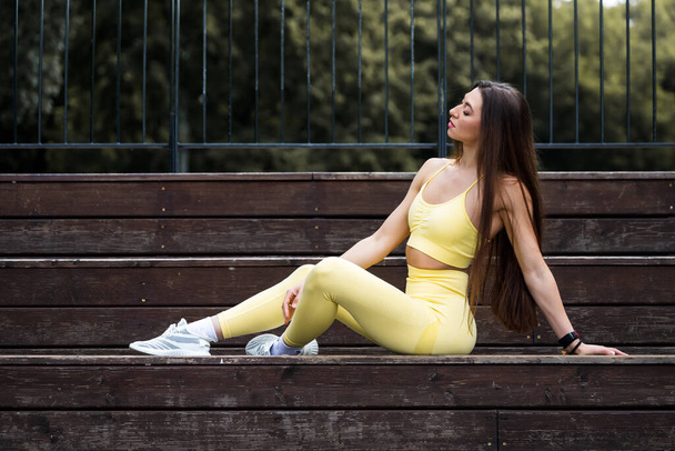 Modeporträt einer jungen Frau mit langen Haaren, in Sportbekleidung, die auf einer Bank in einem Stadtpark in der Sommerstraße sitzt. Porträt einer lebensfrohen Frau - Foto, Bild