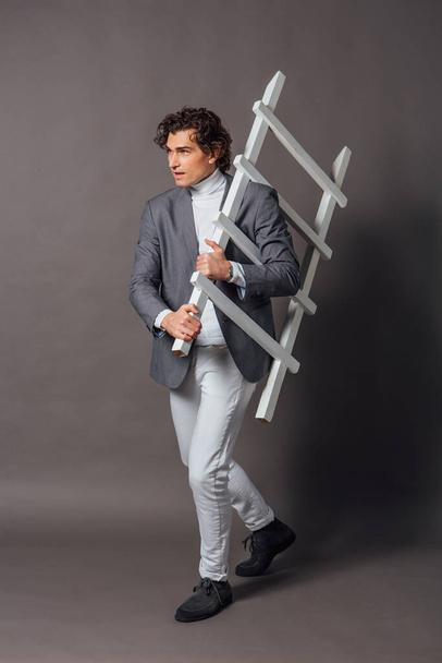 Fashion portrait of a tall handsome man dressed in white turtleneck, white jeans and grey jacket posing on the grey background with white ladder - Zdjęcie, obraz