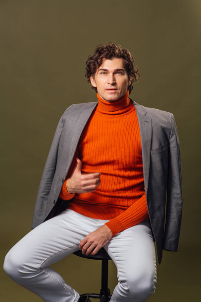Portrait of a tall handsome man dressed in orange turtleneck, grey jacket and white jeans posing on the green background - Foto, Imagen
