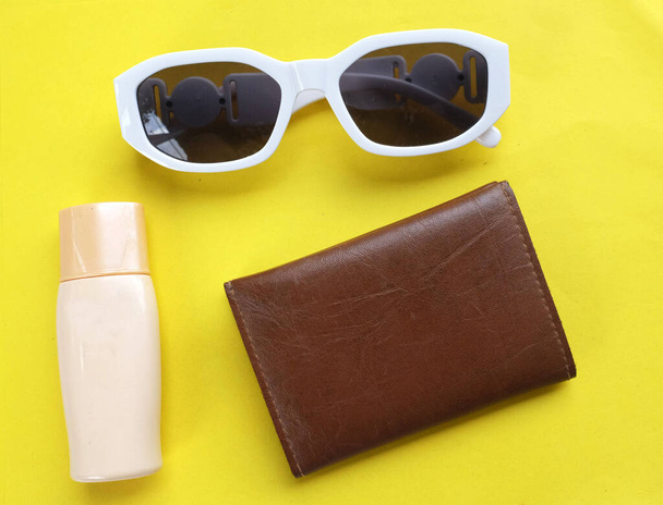 top view and flat lay Sunscreen and white sunglasses The brown purse is on a yellow background. Supplies for a summer trip to the beach. - Photo, Image