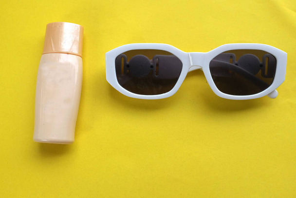  Плоский лежал солнцезащитный крем и белые солнцезащитные очки на желтом фоне. Припасы для летней поездки на пляж. - Фото, изображение