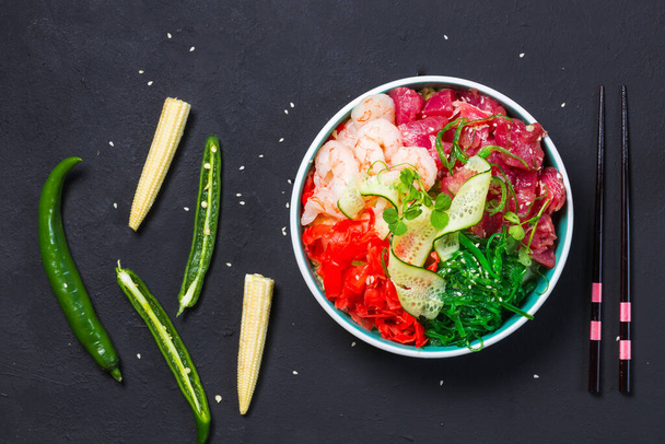 Poke with tuna, shrimp and pike in a round bowl on a black background with chopsticks, mini corn and green hot pepper. Top view, horizontal - Фото, изображение
