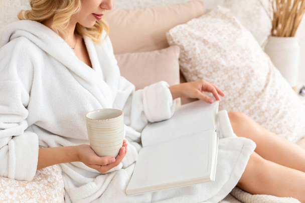 Νεαρή γυναίκα σε λευκό terry ρόμπα πίνει καφέ και διαβάζοντας περιοδικό ή βιβλίο στην κρεβατοκάμαρα. - Φωτογραφία, εικόνα