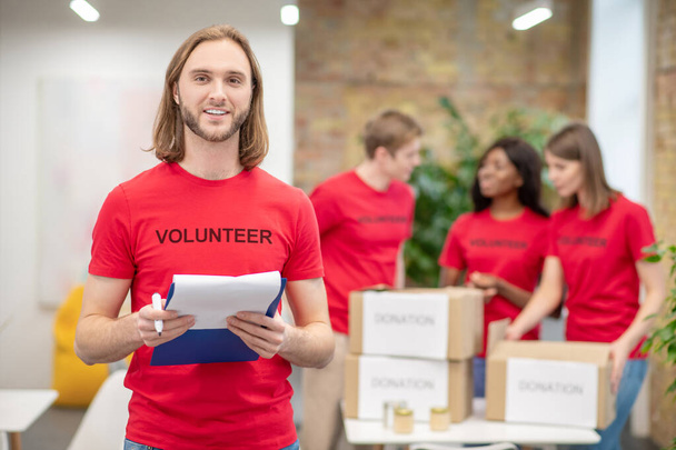 Guy in volunteer tshirt with document and colleagues behind - Photo, image