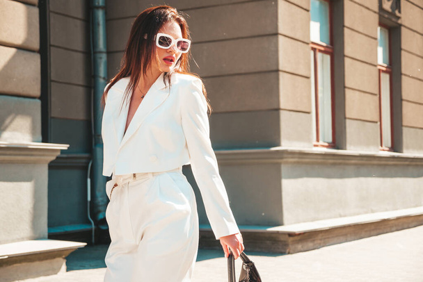 Portrait of young sexy modern businesswoman model. Hot beautiful woman in white suit posing on the street background. Fashion female walking outdoors in sunglasses - Photo, image