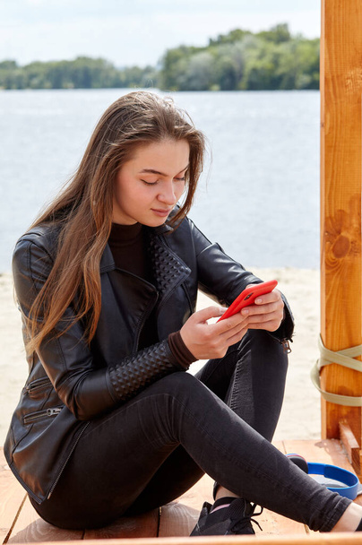 Young caucasian woman is typing on the smartphone while sitting near the river bank. Portrait photo of happy hipster girl in common clothes relaxing at nature and have a good time - Φωτογραφία, εικόνα