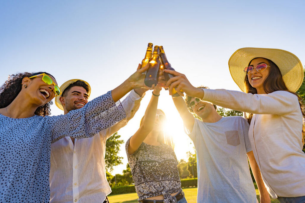 Группа молодых хипстерских друзей, празднующих пиво на открытом воздухе и тосты с бутылкой перед закатом солнца. Счастливые миллениалы веселятся с алкоголем на свежем воздухе в парке в сумерках - Фото, изображение