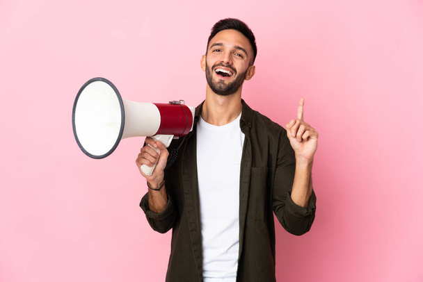 Young caucasian man isolated on pink background holding a megaphone and pointing up a great idea - Photo, Image