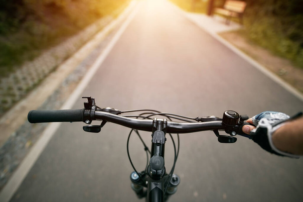 Άποψη πρώτου προσώπου του χειρισμού του ποδηλάτου στον άδειο ασφαλτοστρωμένο δρόμο της πόλης προς το φως του ήλιου - Φωτογραφία, εικόνα