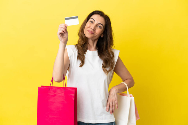 Middle aged caucasian woman isolated on yellow background holding shopping bags and a credit card - Fotografie, Obrázek