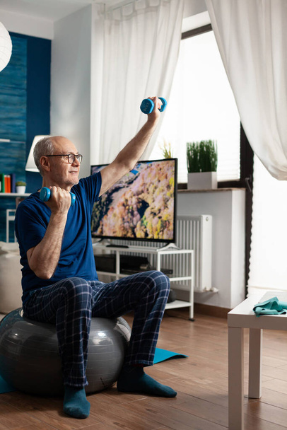 Старший чоловік, який сидить на аеробіці, піднімає м'яч руки, розтягуючи м'язи рук
 - Фото, зображення