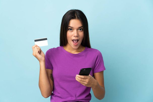 Genç kız pembe arka planda izole edilmiş cep telefonuyla alışveriş yapıyor ve elinde şaşırmış bir ifadeyle bir kredi kartı tutuyor. - Fotoğraf, Görsel
