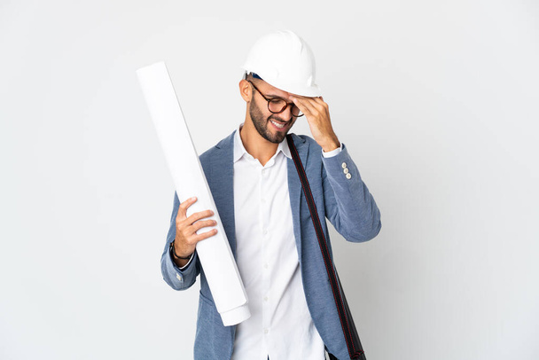 Mladý architekt muž s helmou a drží plány izolované na bílém pozadí s bolestí hlavy - Fotografie, Obrázek