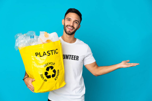 Jonge blanke man die een zak vol plastic flessen vasthoudt om te recyclen geïsoleerd op blauwe achtergrond en handen naar de zijkant strekt om uit te nodigen om te komen - Foto, afbeelding