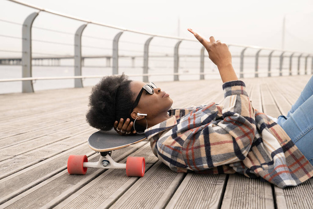 Zamyšlená africká žena s chytrým telefonem vážné ležící na skateboardu na městském nábřeží městský prostor nevrlý - Fotografie, Obrázek
