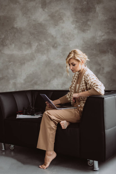 Jonge mooie vrouw blogger in een modern loft interieur. Vrouwelijke freelance fotograaf in beige kleding werkt op een laptop. Zachte selectieve focus. - Foto, afbeelding