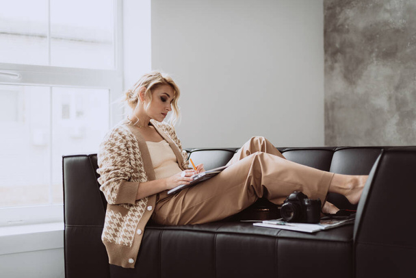 Jonge invloedrijke vrouwelijke blogger werkt op afstand thuis in een modern loft interieur. Vrouwelijke freelance fotograaf in beige kleding. Zachte selectieve focus. - Foto, afbeelding