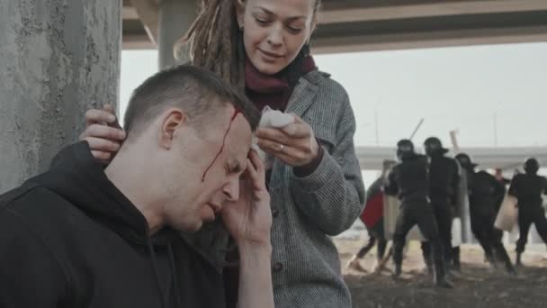 Close up slow-shot di giovane donna con dreadlocks aiutare l'uomo ferito mentre la polizia antisommossa respinge i manifestanti in background - Filmati, video