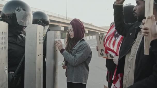 Slowmo śledzenia ujęcia młodej kobiety z dredami wprowadzenie kwiatów na tarcze policji zamieszek podczas gdy ludzie ze znakami i USA flaga protestują - Materiał filmowy, wideo