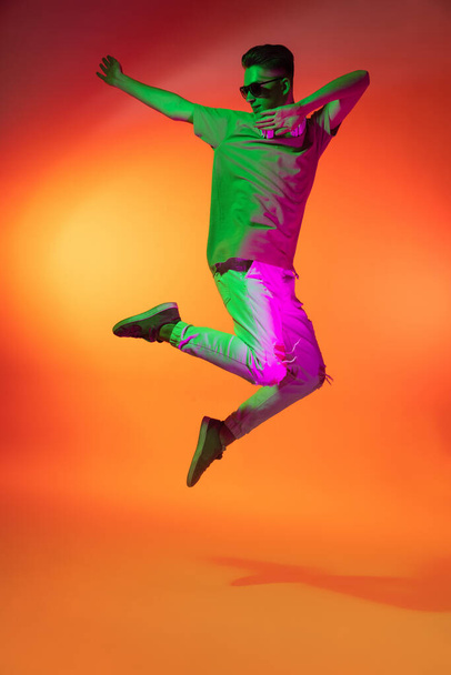 Portrait of Caucasian young man jumping, dancing isolated on orange studio background in neon light. Concept of human emotions, facial expression. - Foto, Bild