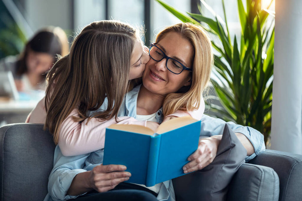 Foto de mujer madura hermosa leyendo un libro mientras su hija abrazándose en la sala de estar en casa. - Foto, Imagen