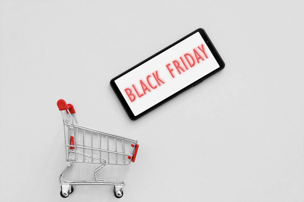 Shopping cart with a smartphone and text Black Friday on the screen on a grey background. Season of sales and concept of an online store. - Φωτογραφία, εικόνα