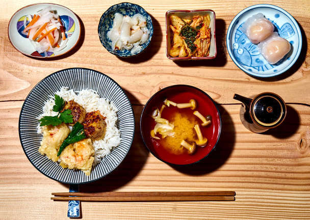 A top view of bowls with grilled scallops, crab tempura on rice, miso soup with mushrooms, radish, kimchi, steamed dumplings, and lychee - Фото, зображення