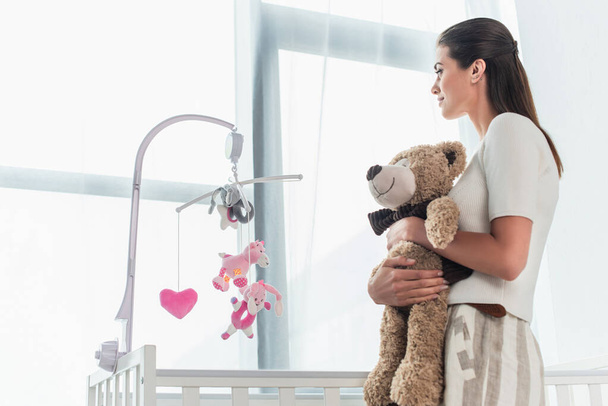 Vue latérale de la femme tenant un ours en peluche près du lit de bébé à la maison  - Photo, image