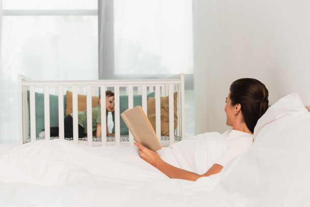 Woman with book lying on bed near child in baby bed on blurred background  - Foto, Bild