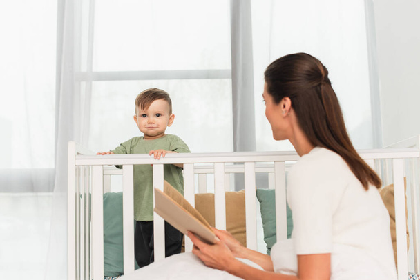 Kid standing in baby bed near blurred mother with book  - Fotó, kép