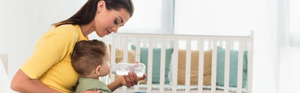 Young mother holding bottle of water near child at home, banner  - Фото, изображение