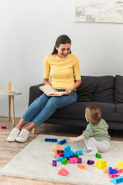Happy mother holding book near kid with building blocks on carpet  - Valokuva, kuva