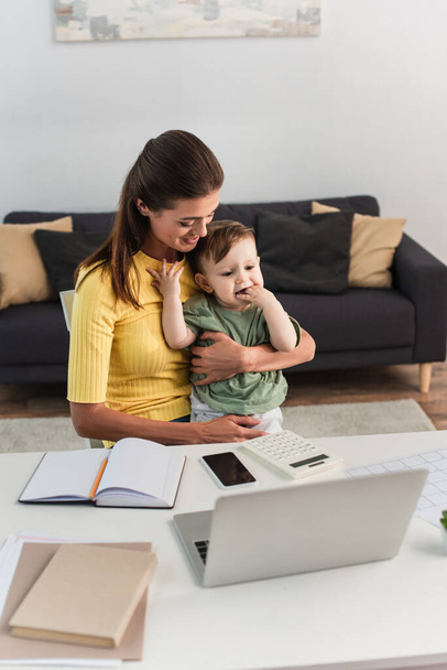 Smiling freelancer hugging son near devices and notebooks at home  - Foto, Bild