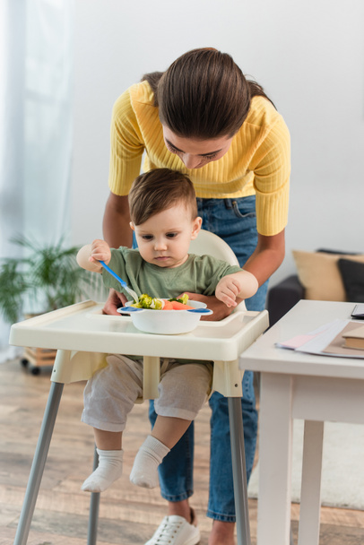 Mother standing near kid and food on high chair  - Foto, immagini