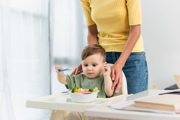 Woman hugging child with spoon near bowl of food on high chair  - Foto, afbeelding