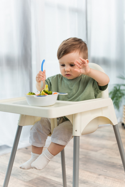 Toddler boy holding spoon near blurred bowl of vegetables on high chair  - Foto, Imagen