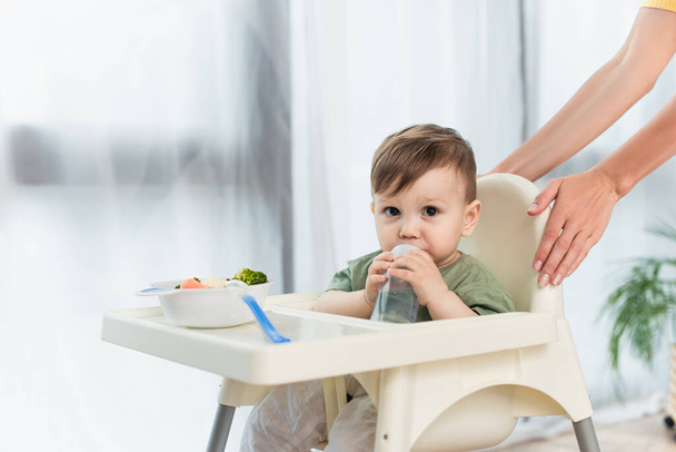 Woman standing near toddler son with baby bottle and food on high chair  - Fotó, kép