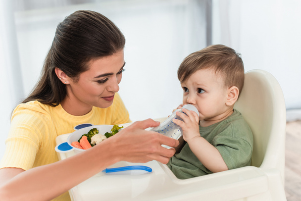Mother holding baby bottle near son and vegetables on high chair at home  - Foto, Imagem
