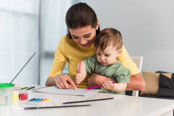 Young woman smiling while pointing at paper near son with pencils at home  - Foto, Imagen