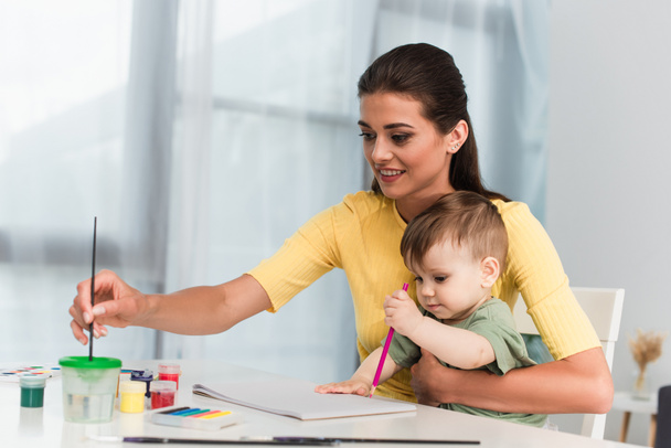 Smiling woman holding paint brush near child with pencil  - Fotó, kép